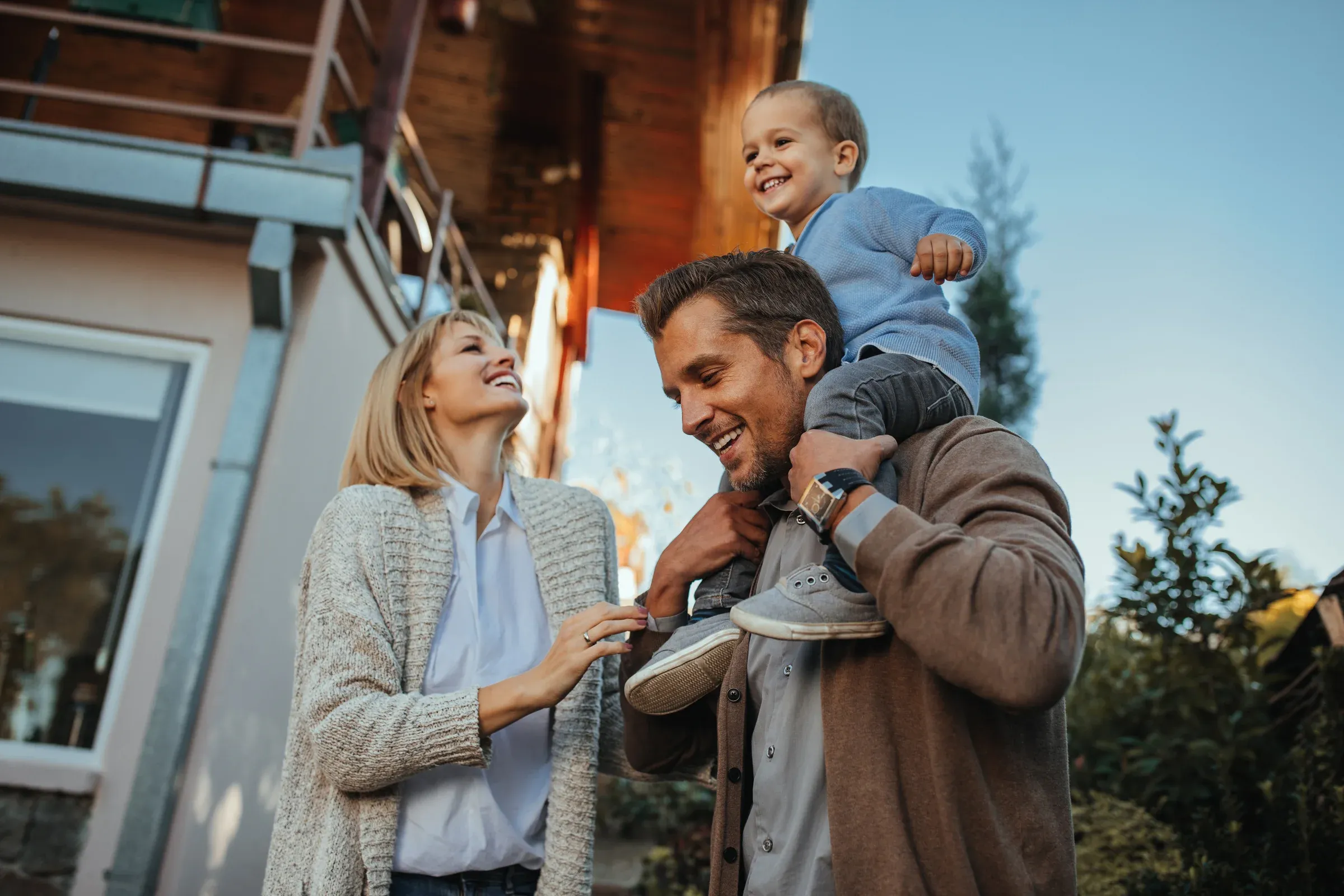 Familie lacht gemeinsam vor ihrem Haus, der Vater trägt das Kind auf den Schultern